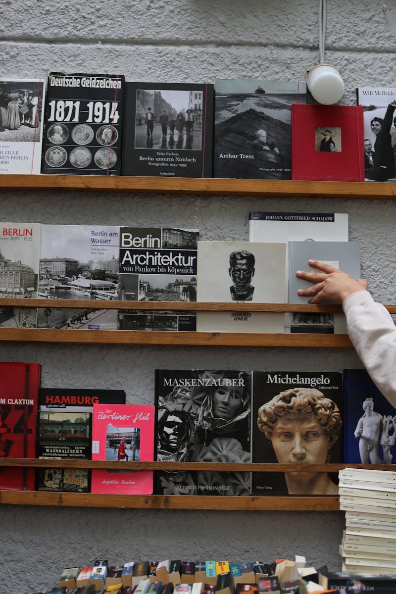 A diverse collection of books on a wooden shelf in a Berlin bookstore, with a hand browsing them.