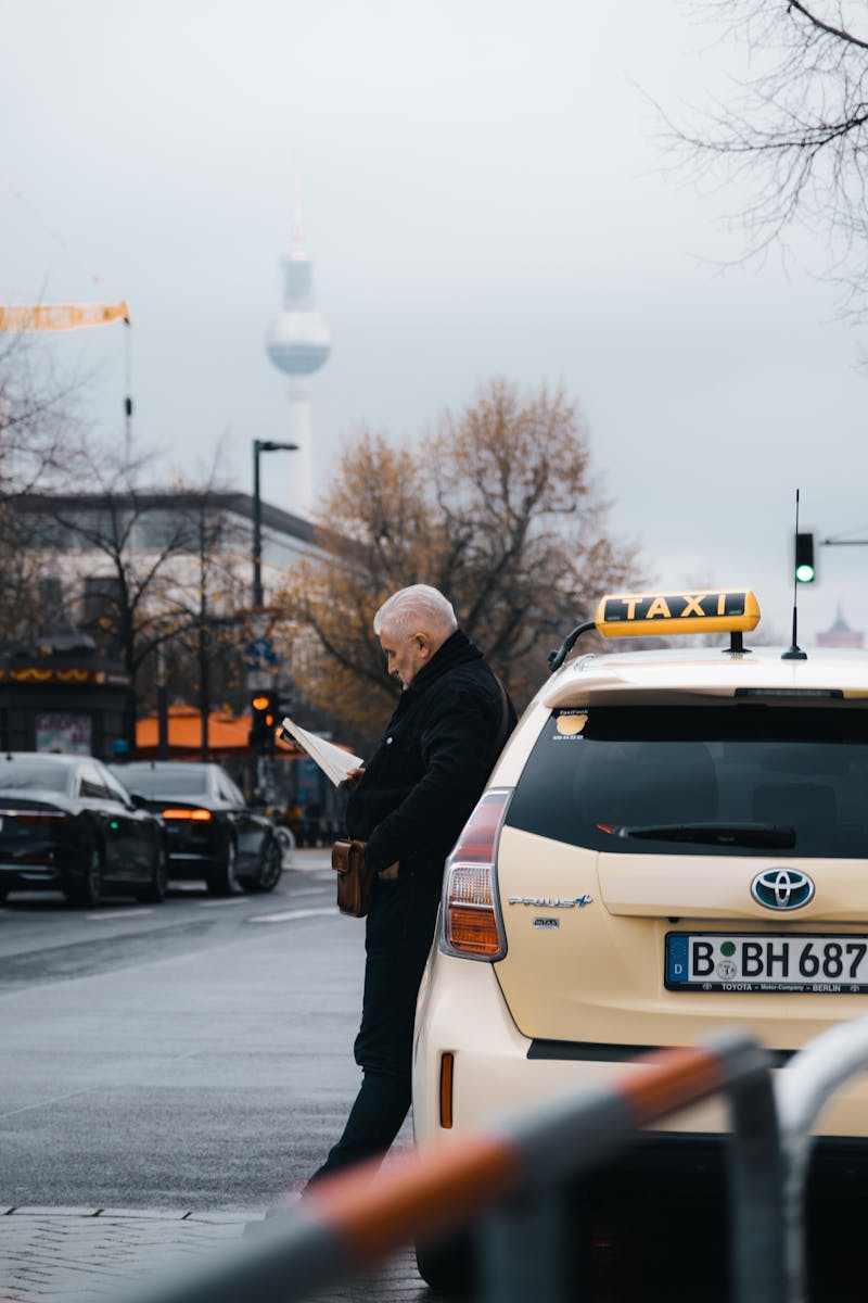 An elderly man reads beside a taxi on a city street in Berlin, Germany, with the TV Tower in the background.
