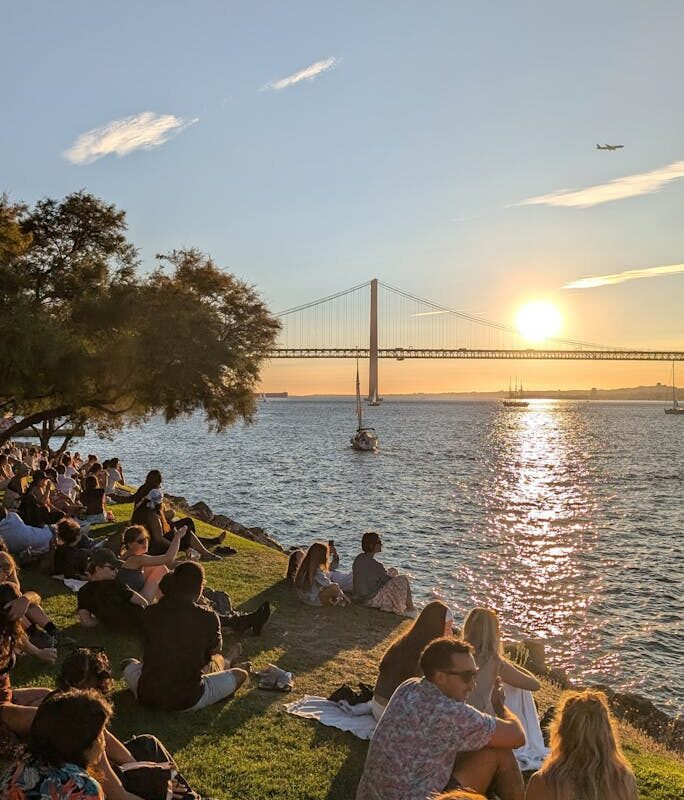 A vibrant scene of people relaxing by the Tagus River in Lisbon at sunset with a view of the suspension bridge.