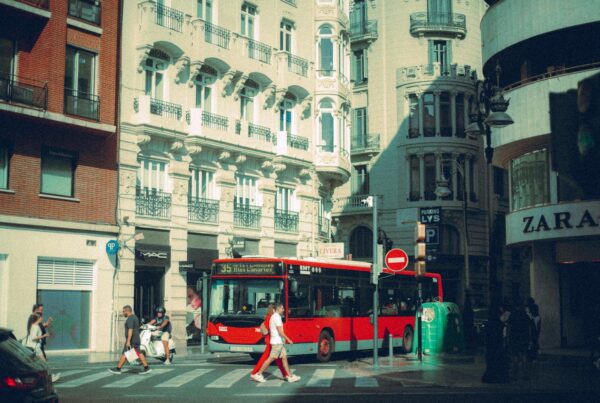 Red city bus at busy crosswalk in front of ornate historic buildings.
