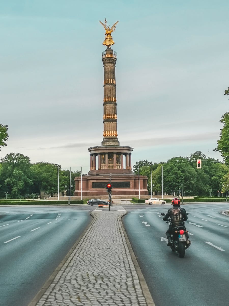 A motorcyclist rides towards Berlin's iconic Victory Column on a tree-lined avenue.