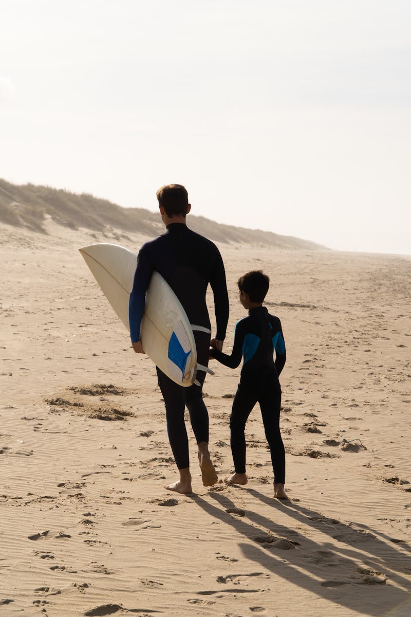 A father and son walk on a sandy beach in Portugal, carrying a surfboard, enjoying quality time together.