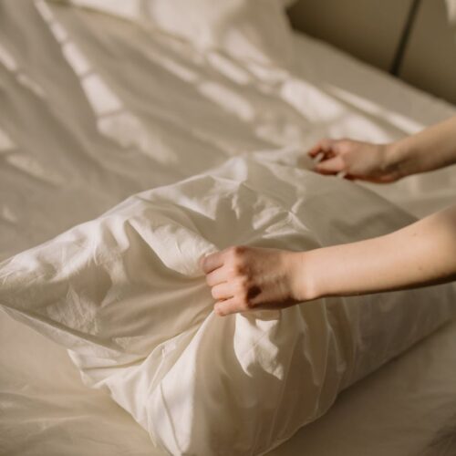 A close-up of hands adjusting a pillow on a neatly made bed, bathed in warm sunlight.